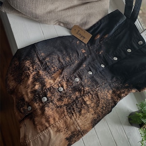 May include: A black denim overall dress with a bleach-splattered design. The dress has a button closure and pockets. The dress is handmade by "Artsyguy".