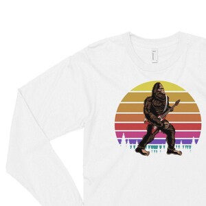 May include: White long-sleeve t-shirt with a graphic of a Bigfoot playing an electric guitar in front of a sunset. The graphic is in a vintage style with a rainbow color scheme.