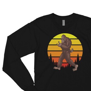 Puede incluir: Camiseta negra de manga larga con un gráfico de Bigfoot de pie frente a una puesta de sol. El gráfico es de estilo retro con una puesta de sol amarilla, naranja y roja.