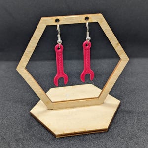 Puede incluir: Un par de pendientes de llave inglesa roja en un expositor de madera. Los pendientes son de plástico y tienen un gancho plateado.