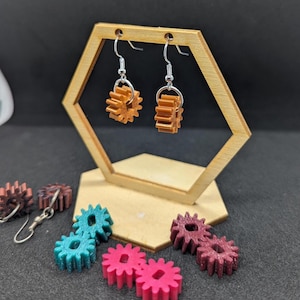 Puede incluir: Un par de pendientes marrones con forma de engranaje y ganchos plateados se exhiben en un soporte hexagonal de madera. Varios objetos coloridos con forma de engranaje en color turquesa, rosa y granate están dispersos.