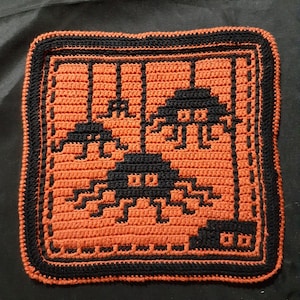 May include: A crocheted square pillow with a black border and an orange center. The center features a pixelated design of three black space invaders.