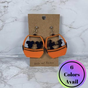 May include: A pair of orange acrylic earrings with black character silhouettes inside a gondola design. The earrings are on a brown card with "Vector and Romance" text. A circular graphic states "6 Colors Avail".