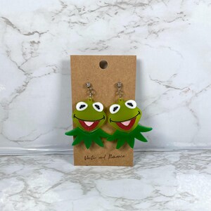 May include: A pair of green frog-shaped earrings with white eyes and red mouths. The earrings are attached to silver-coloured posts and hang from a brown card. The card has the text "Vector and Romance" printed on it.