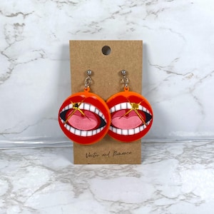 May include: A pair of orange and red acrylic earrings with a cartoon mouth design. The earrings are round with a mouth graphic, black teeth, and a pink tongue. They are attached to silver posts and displayed on a brown card. The text "Vector and Romance" is visible.