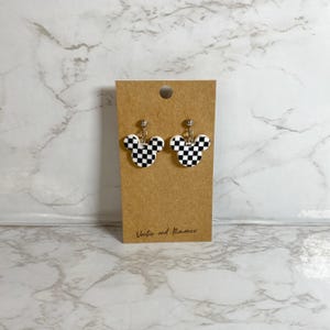 May include: A pair of earrings shaped like Mickey Mouse heads, featuring a black and white checkered pattern. The earrings are attached to silver-colored posts and are displayed on a brown card. The card has the text "Vector and Romance" printed on it.