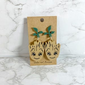 May include: A pair of green and brown earrings featuring a cartoon character with a smiling face and leaves on top. The earrings are displayed on a brown card with the text "Victor and Romance".