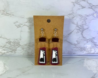 May include: A pair of red llama earrings with white and black details. The earrings are on a brown card.