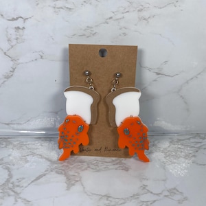 May include: A pair of novelty earrings featuring a slice of white bread with a brown crust, topped with an orange fish-shaped design with blue spots. The earrings are attached to a brown card with the text "Vector and Romance".