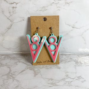 May include: A pair of turquoise and pink earrings with a V-shaped design. The earrings are made of acrylic and have a silver hook closure. The earrings are labeled "Vector and Roman".