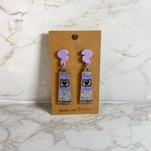 May include: A pair of purple and silver glitter tube earrings with a black Mickey Mouse head on each tube. The earrings are hanging from a purple cloud-shaped charm.