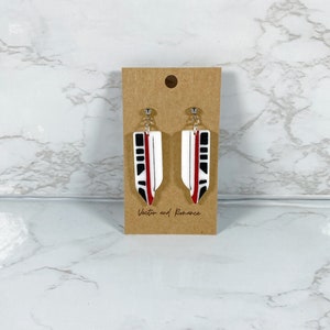 Peut inclure: Une paire de boucles d'oreilles pendantes blanches, noires et rouges. Chaque boucle d'oreille a une forme rectangulaire avec un motif noir et rouge. Les boucles d'oreilles sont fixées à une tige de couleur argentée. Les boucles d'oreilles sont présentées sur une carte marron avec le texte "Vector and Romance".