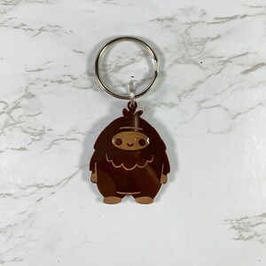 May include: A brown, cartoon-style yeti keychain with a smiling face. The keychain is attached to a silver key ring.