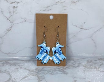 May include: A pair of dangle earrings featuring a light blue and white design. The earrings are shaped like stylized figures with a geometric pattern. They hang from silver-colored ear wires and are displayed on a brown card.