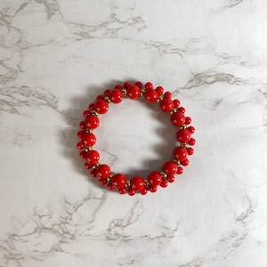 May include: A red beaded bracelet with a Mickey Mouse-inspired design. The bracelet features round red beads and smaller red beads with gold-colored accents. The bracelet is displayed on a white and gray marble surface.