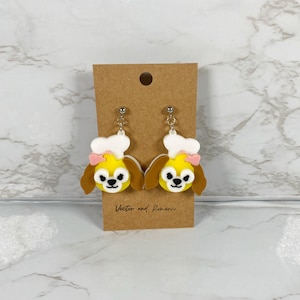 May include: A pair of dangle earrings featuring a cartoon dog wearing a chef's hat. The dog is yellow with brown ears and a pink bow. The earrings are made of polymer clay and have silver hooks.
