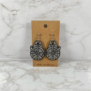 May include: A pair of silver glitter earrings shaped like the Millennium Falcon from Star Wars. The earrings are on a brown card with the text "Vector and Romance".