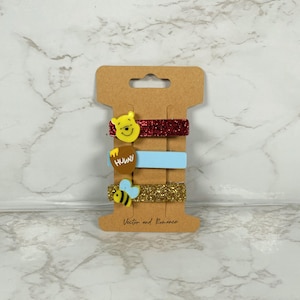 May include: A set of four hair clips on a brown cardboard backing. The clips feature a yellow Winnie the Pooh, a red glitter clip, a light blue clip, a honey pot, a gold glitter clip, and a bee. The text "Vector and Romance" is printed on the bottom.
