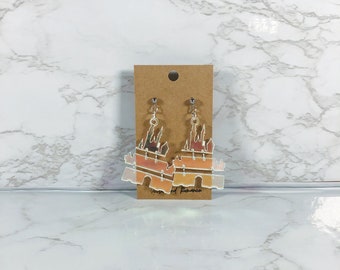 May include: A pair of dangle earrings featuring a castle silhouette design with a gradient of orange, peach, and white colours. The earrings are attached to a brown rectangular display card with a small hole at the top for hanging purposes.