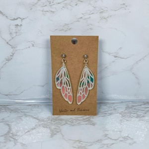 May include: Silver-coloured butterfly wing earrings with pink and green details. The earrings are presented on a brown card with the text "Vector and Romance". A distinctive accessory.