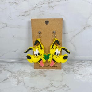 May include: A pair of yellow Pluto dog earrings with black ears and a pink tongue. The earrings are attached to a brown card with the text "Vector and romance". The earrings are hanging from silver-colored hooks.