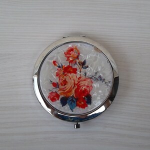 May include: A silver compact mirror with a floral design. The design features red, orange, and yellow roses on a white background.