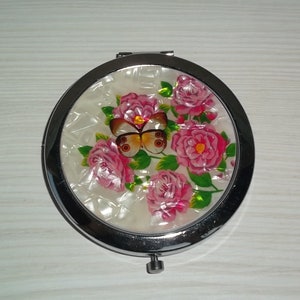 May include: A round, silver-framed compact mirror with a white background and a pink and green floral design featuring a butterfly.