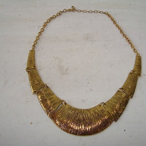 May include: A gold-toned statement necklace with a textured, curved design. The necklace has a chain that connects to the front piece.