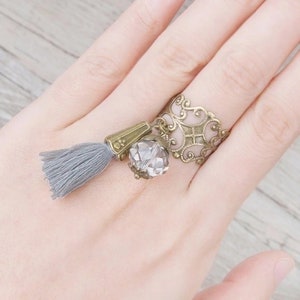 May include: A silver filigree ring with a crystal bead and a gray tassel hanging from it.