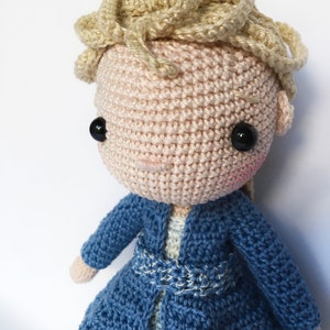 K&ouml;nnte beinhalten: H&auml;kelpuppe mit blonden Haaren, die ein blaues H&auml;kelkleid und eine blaue H&auml;keljacke tr&auml;gt. Die Puppe hat schwarze Augen und rosige Wangen.