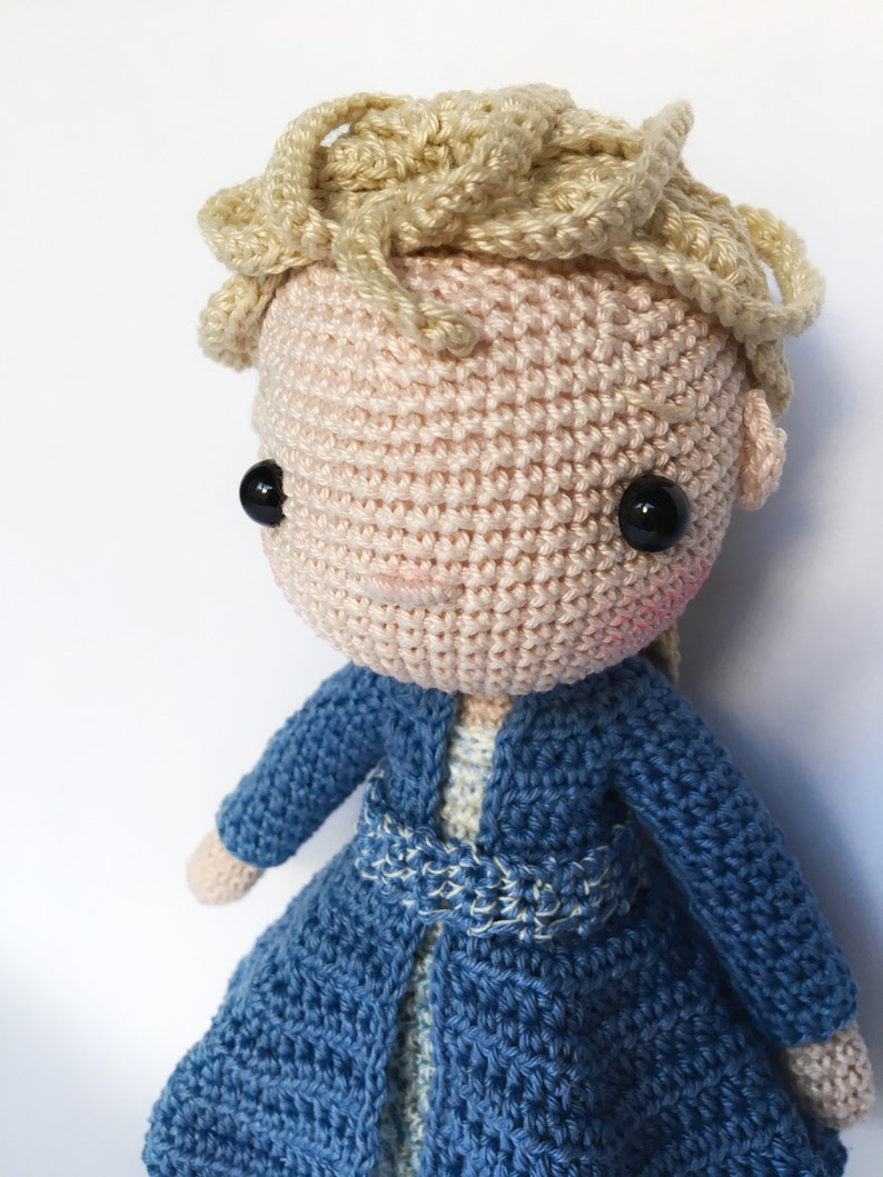 K&ouml;nnte beinhalten: H&auml;kelpuppe mit blonden Haaren, die ein blaues H&auml;kelkleid und eine blaue H&auml;keljacke tr&auml;gt. Die Puppe hat schwarze Augen und rosige Wangen.