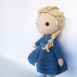 K&ouml;nnte beinhalten: Eine geh&auml;kelte Puppe mit blonden Haaren, einem blauen Kleid und blauen Schuhen. Die Puppe steht auf einem wei&szlig;en Hintergrund.