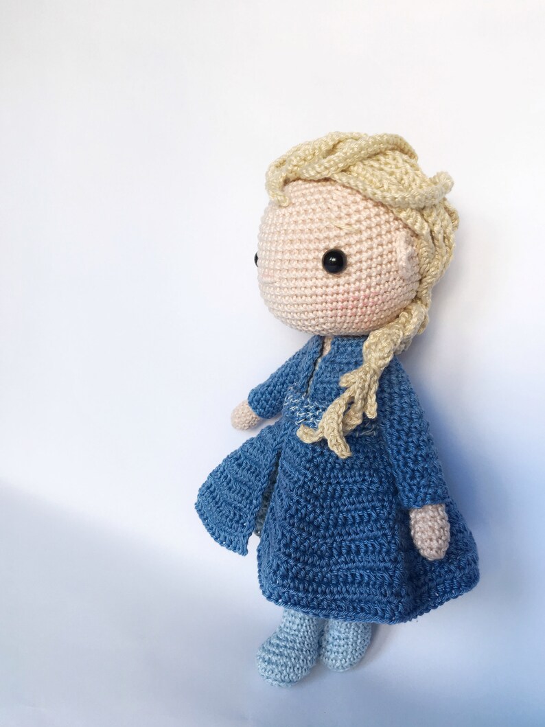 K&ouml;nnte beinhalten: Eine geh&auml;kelte Puppe mit blonden Haaren, einem blauen Kleid und blauen Schuhen. Die Puppe steht auf einem wei&szlig;en Hintergrund.