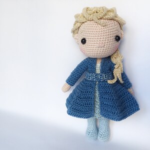 K&ouml;nnte beinhalten: Eine geh&auml;kelte Puppe mit blonden Haaren, die ein blaues Kleid und einen blauen Mantel tr&auml;gt. Die Puppe hat ein rosafarbenes Gesicht und schwarze Augen.