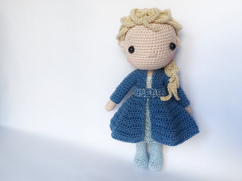 K&ouml;nnte beinhalten: Eine geh&auml;kelte Puppe mit blonden Haaren, die ein blaues Kleid und einen blauen Mantel tr&auml;gt. Die Puppe hat ein rosafarbenes Gesicht und schwarze Augen.