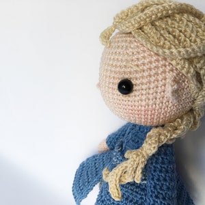 K&ouml;nnte beinhalten: Eine geh&auml;kelte Puppe mit blonden Haaren in einem Zopf, die ein blaues Kleid tr&auml;gt. Die Puppe hat schwarze Augen und einen rosa Teint.
