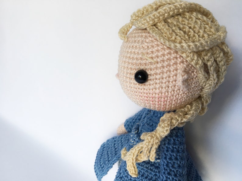 K&ouml;nnte beinhalten: Eine geh&auml;kelte Puppe mit blonden Haaren in einem Zopf, die ein blaues Kleid tr&auml;gt. Die Puppe hat schwarze Augen und einen rosa Teint.
