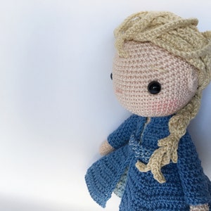 K&ouml;nnte beinhalten: Eine geh&auml;kelte Puppe mit blonden Haaren und einem blauen Kleid. Die Puppe hat einen s&uuml;&szlig;en Ausdruck und tr&auml;gt ein blaues Kleid mit wei&szlig;em Besatz.