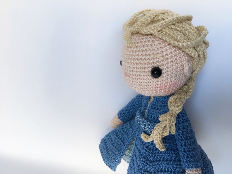 K&ouml;nnte beinhalten: Eine geh&auml;kelte Puppe mit blonden Haaren und einem blauen Kleid. Die Puppe hat einen s&uuml;&szlig;en Ausdruck und tr&auml;gt ein blaues Kleid mit wei&szlig;em Besatz.