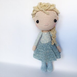 K&ouml;nnte beinhalten: Eine geh&auml;kelte Puppe mit blonden Haaren, einem blau-wei&szlig;en Kleid und schwarzen Augen. Die Puppe steht auf einem wei&szlig;en Hintergrund.