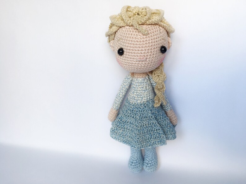 K&ouml;nnte beinhalten: Eine geh&auml;kelte Puppe mit blonden Haaren, einem blau-wei&szlig;en Kleid und schwarzen Augen. Die Puppe steht auf einem wei&szlig;en Hintergrund.