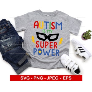 Op de afbeelding: Een grijs T-shirt met de tekst "AUTISM IS MY SUPER POWER" in kleurrijke letters. Boven de tekst bevindt zich een zwart superheldenmasker. Het shirt ligt op een witte achtergrond met een paar witte sneakers en een plant in een pot.
