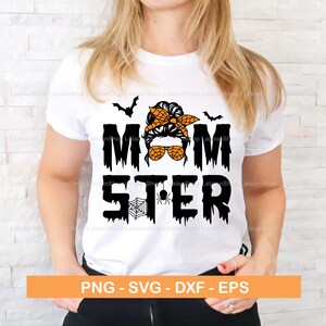 Puede incluir: Una camiseta blanca con un gr&aacute;fico negro y naranja que dice "Momster" con una imagen de dibujos animados de una mujer con un mo&ntilde;o desordenado, gafas de sol y una diadema. El dise&ntilde;o tambi&eacute;n incluye murci&eacute;lagos y una telara&ntilde;a.