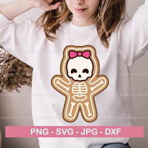 Könnte beinhalten: Ein weißes Sweatshirt mit einem Lebkuchen-Skelett-Design und einer rosa Schleife. Das Lebkuchen-Skelett hat ein Totenkopfgesicht und einen sichtbaren Brustkorb. Der Text "PNG - SVG - JPG - DXF" ist enthalten.