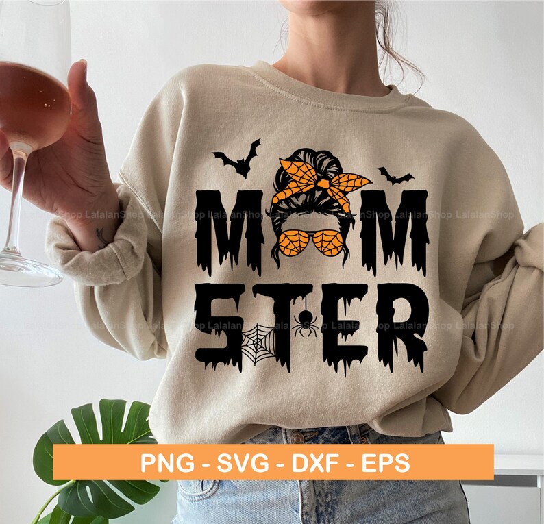 Puede incluir: Una sudadera beige con un dise&ntilde;o gr&aacute;fico negro que dice "Momster" con una ara&ntilde;a negra y una telara&ntilde;a. El gr&aacute;fico tambi&eacute;n incluye una imagen de dibujos animados de una mujer con un mo&ntilde;o desordenado, gafas de sol y un pa&ntilde;uelo. El texto gotea con un l&iacute;quido negro.
