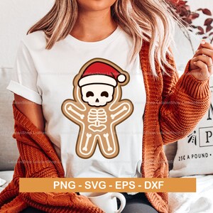 以下が含まれることがあります： 白いTシャツにジンジャーブレッドマンのデザインがプリントされています。ジンジャーブレッドマンは赤いサンタの帽子をかぶり、骨格のデザインになっています。デザインの下には「PNG - SVG - EPS - DXF」というテキストが印刷されています。