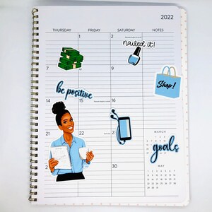 May include: A monthly planner with stickers featuring a woman with a blue shirt and a book, a stack of money, a bottle of nail polish, a shopping bag, and a phone with headphones. The planner has the words "nailed it!" and "Shop!" written on it. The planner also has the words "be positive" and "goals" written on it.