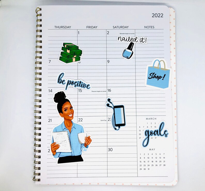 May include: A monthly planner with stickers featuring a woman with a blue shirt and a book, a stack of money, a bottle of nail polish, a shopping bag, and a phone with headphones. The planner has the words "nailed it!" and "Shop!" written on it. The planner also has the words "be positive" and "goals" written on it.