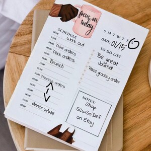 May include: A white planner with a pink and brown illustration of a hand holding a coffee mug. The planner has a daily schedule, a to-do list, and notes section. The text "focus on today" is written in cursive on the top of the planner. The date is 01/15.