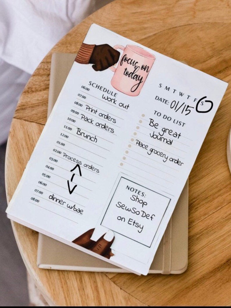 May include: A white planner with a pink and brown illustration of a hand holding a coffee mug. The planner has a daily schedule, a to-do list, and notes section. The text "focus on today" is written in cursive on the top of the planner. The date is 01/15.